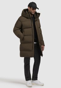 Brauner Puffermantel mit Kapuze, mit durchgehender Knopfleiste und Seitentaschen. Tragen über einem schwarzen Pullover und kombiniert mit dunklen Jeans und weißen Sneakern.