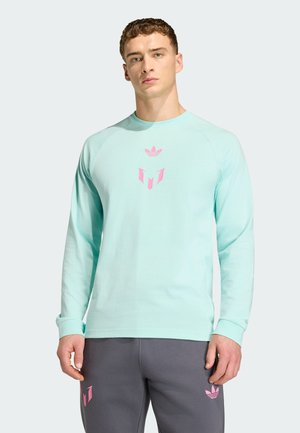 Mann trägt hellblaues Langarmhemd mit pinken Logos und graue Jogginghose mit pinken Logos, steht vor einem schlichten, hellen Hintergrund.