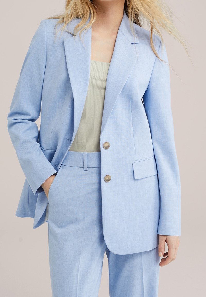 Femme portant un costume bleu clair sur mesure avec une veste et un pantalon assortis, associé à un top beige côtelé, main dans la poche.
