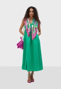 Grön, ärmlös maxiklänning med en utsvängd kjol, kombinerad med en mönstrad rosa scarf och en volangförsedd rosa handväska. Remsandaler i rosa.