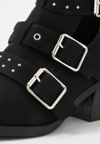 Stivale alla caviglia in suede nero con fibbie argentate, dettagli borchiati e tacco impilato. Presenta un design a tracolla e una superficie testurizzata.