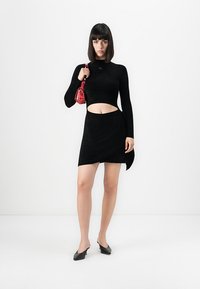 Žena so rovnými tmavými vlasmi, oblečená v čiernom topi s vykrojením a sukni, drží malú červenú kabelku, stojí na bielom pozadí.