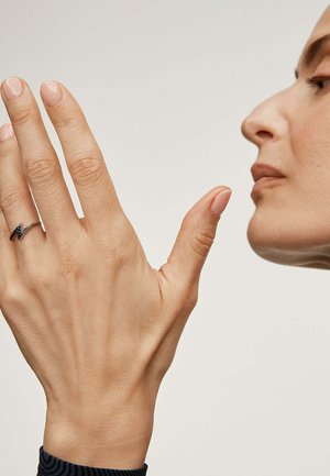 Anillo de plata con un diseño en zigzag, con pequeños acentos negros. Mano mostrada con esmalte de uñas neutro contra un fondo claro.