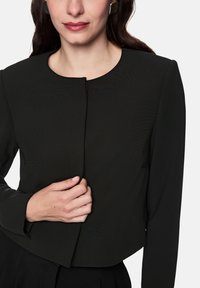 Black cropped blazer with a round neckline, long sleeves, tailored design, and a single front closure. Smooth texture, minimal detailing.
