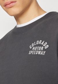 Šedá mikina s žebrovaným límcem, na hrudi s bílým vyšitým logem "COLORADO MOTOR SPEEDWAY". Hladká struktura látky.