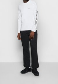 Person, der ein weißes Langarmshirt mit "Calvin Klein"-Logo, schwarze Hosen, weiße Socken und schwarze Turnschuhe trägt, steht vor einem schlichten Hintergrund.