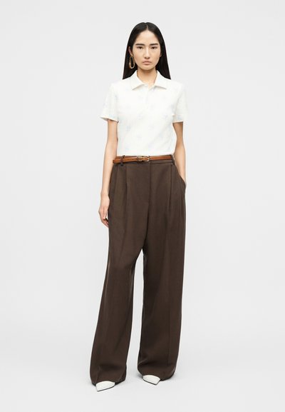 Femme debout les mains dans les poches, portant une chemise blanche à manches courtes, un pantalon marron large à ceinture, et des chaussures blanches pointues.