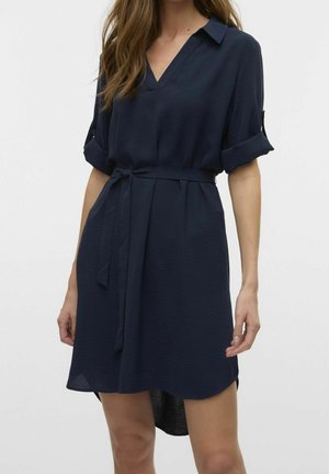 Femme portant une courte robe bleu marine avec des manches retroussées, un col en V et une ceinture nouée à la taille.