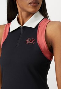 Detailní záběr ženy v bezramenném černém a červeném topu s bílým límcem a logem EA7 Emporio Armani na hrudi.