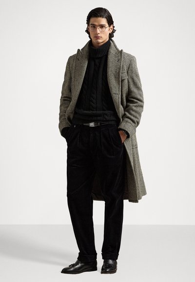 Abrigo de espiga gris, cuello de tortuga negro, pantalones de pana negros, mocasines. Gafas puestas; manos en los bolsillos, pose de pie, fondo minimalista.