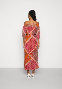 Kvinna med flätat hår som bär en av-axel lång mönstrad klänning i röd, orange och vit, stående vänd mot en vit vägg.