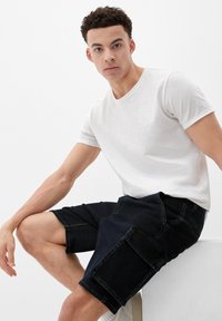 Dunkelblaue Cargo-Shorts aus Denim mit mehreren Taschen, kombiniert mit einem schlichten weißen T-Shirt. Das Material wirkt weich und leicht.