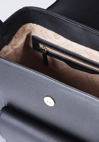 Mustan nahkakäsilaukun sisäosa, jossa on beige kangaspäällyste ja tekstiä painettuna. Laukussa on myös pieni vetoketjullinen tasku, jossa on kultasävyinen vetoketjun vedin.