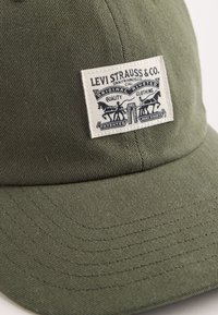 Olijfgroene baseballcap van katoen, met een rechthoekig wit logopatch met zwarte tekst en paardenafbeeldingen.