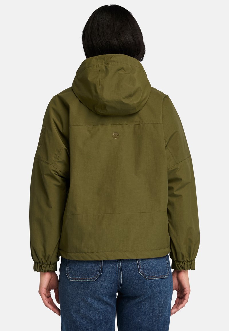 Giacca a vento verde oliva con cappuccio, polsini elasticizzati, una consistenza liscia e un dettaglio del logo sulla schiena, abbinata a jeans in denim blu.