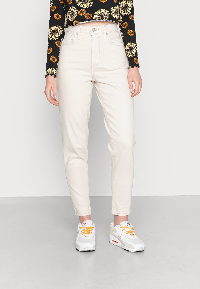 Femme portant un jean taille haute blanc cassé, un haut noir à manches longues avec un imprimé floral jaune et des baskets blanches avec des lacets orange.