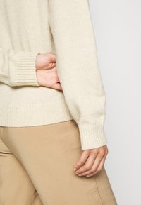 Beige stickad tröja med ribbade ärm- och nederkant, som visar en mjuk textur. Handen vilar nära fickområdet, i kombination med ljusbeige byxor.