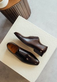 Chaussures habillées en cuir marron avec une finition lisse, un bout pointu et un système de laçage, posées sur une surface blanche texturée.
