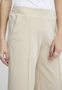 Beige vide bukser laget av teksturert stoff, med sidelommer og en plisserte midje med tre horisontale sømmer.
