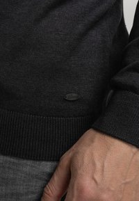 Ciemnoszary sweter z delikatnej dzianiny z prążkowanymi mankietami i dołem, z małą czarną metką na boku.