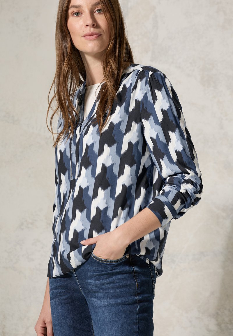 Blusa leggera e a fantasia caratterizzata da colori blu, nero e bianco. Maniche lunghe con polsini arrotolati; sottili pieghe sulle spalle.