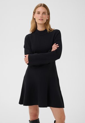 Femme aux cheveux blonds lisses portant une robe noire à manches longues et des bottes noires, debout les bras croisés devant un fond uni.