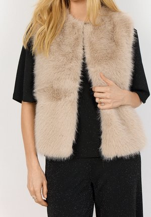 Blonde vrouw draagt een beige, pluizig mouwloos vest over een zwart glinsterend shirt met korte mouwen en bijpassende broek, houdt het voorpand van het vest vast.