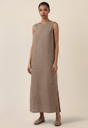 Kvinde iført ærmeløs, ankellang taupe linned kjole med knapper i siden og slids, kombineret med brune flade sandaler med remme.