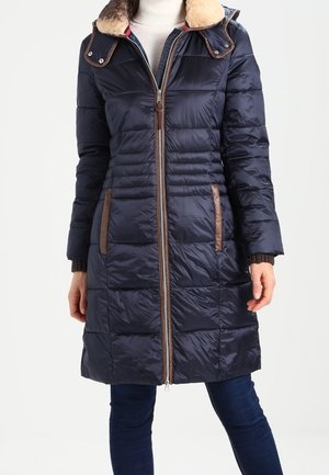 Kobieta ubrana w granatowy pikowany długi puchowy płaszcz z brązowymi lamówkami i futrzanym kołnierzem, na ciemnych dżinsach.