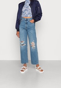 Giacca bomber blu navy, top corto floreale, jeans svasati chiari e strappati, e sandali bianchi con cinturini. Outfit semplice e casual.