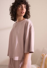 Maglietta loose-fitting rosa chiaro con scollatura rotonda e maniche a lunghezza gomito, realizzata in tessuto morbido con una texture liscia, con dettagli hardware minimi.