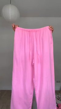 Χέρια κρατούν φαρδιά ροζ παντελόνια με κορδόνι στη μέση σε ένα μινιμαλιστικό δωμάτιο με γκρι τοίχους και ξύλινο πάτωμα.