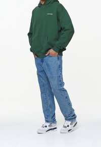 Vihreä huppari taskulla ja logolla, yhdistetty vaaleansinisiin farkkuihin ja valkoisiin lenkkikengiin mustilla yksityiskohdilla. Rentoa tyyliä rennossa istuvuudessa.