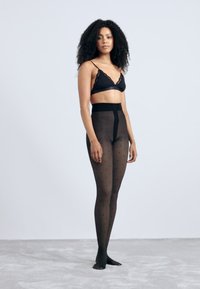 Black opaque tights with a high waist and polka dot pattern, made from a smooth, stretchy material, complementing a black lacy bra.