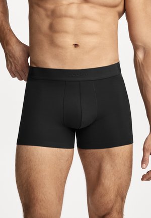 Männlicher Torso mit enganliegenden schwarzen Boxershorts mit sichtbarem Jockey-Logo auf dem Bund vor einfarbigem Hintergrund.