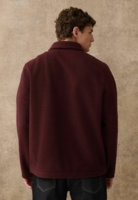 Bordeauxroter Fleece-Pullover mit strukturierter Oberfläche, einem Kragen und langen Ärmeln. Der Saum ist gerade und wird mit dunklen Jeans kombiniert.