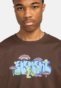 Bruine katoenen T-shirt met een kleurrijk grafisch ontwerp, met blauwe tekst en een cartoonachtige groene worm tussen gestileerde oranje paddenstoelen.