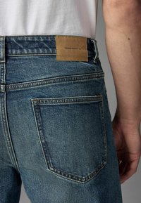 Jeans din denim cu un finisaj albastru închis, având un brat din piele maro etichetat "DENIM SUPPLY Co." la talie și detalii subtile de cusătură.