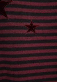 Luumu- ja mustaraidallinen kangas, jossa kaksi samettista punaista tähteä, toinen suurempi kuin toinen, lisäten tekstuuria ja visuaalista kiinnostavuutta.