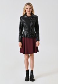 Giacca di pelle nera con un fronte arricciato e orlo peplum, abbinata a una gonna svasata bordeaux e stivali neri alla caviglia. Sfondo semplice.