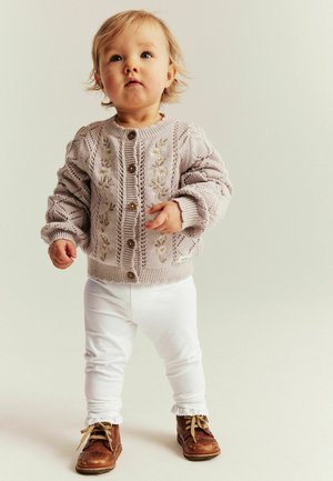 Gestrickte Strickjacke in sanftem Lavendel mit Blumenstickerei, braunen Holzknöpfen, dazu weiße Hosen und braune Schnürstiefel.