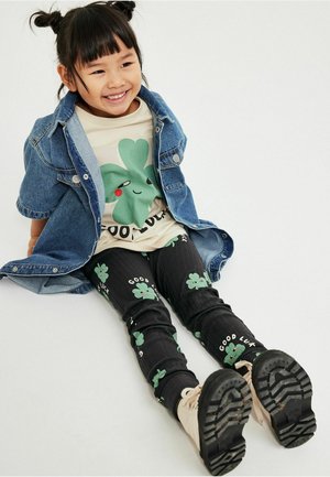 Lächendes Kind sitzt mit ausgestreckten Beinen, trägt eine Jeansjacke, ein cremefarbenes Shirt mit grünem Kleeblatt, schwarze Hosen mit Kleeblatt- und "Good luck"-Text sowie beige Stiefel.