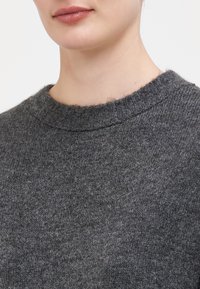 Grå ullgenser med ribbestrikket hals, myk tekstur og klassisk rund halslinning, vist fra skuldrene og opp mot en hvit bakgrunn.