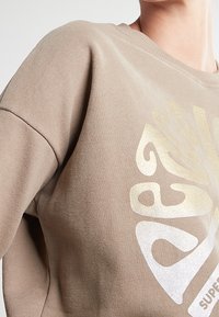 Sudadera beige con letras abstractas metálicas en dorado y plateado en el pecho, llevada por una persona mostrada desde el cuello hasta la mitad del torso.