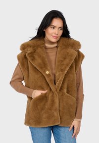 Donna con lunghi capelli scuri che indossa un gilet senza maniche in finta pelliccia marrone sopra un maglione a collo alto marrone e jeans blu, con una mano in tasca.
