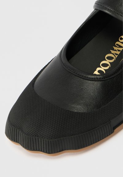 Melnas kurpes ar teksturētu gumijas purngalu un zoli, gluda ādas augšdaļa un zelta teksts, kas redzams uz ieliekamā daļa.