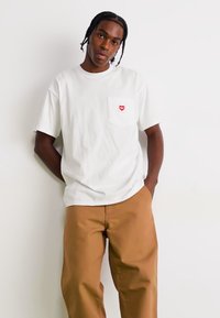 Un tricou alb din bumbac, cu un buzunar și un emblema în formă de inimă roșie, purtat cu pantaloni maroni, sprijinit de un zid.