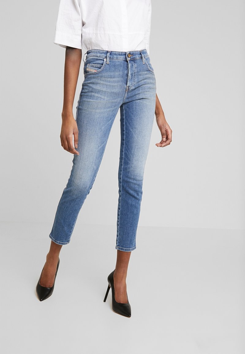 Femme portant un jean slim bleu clair, un haut blanc et des chaussures à talons noirs, debout devant un fond gris uni.