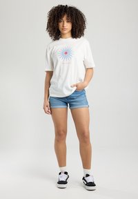 Maglietta bianca oversize con grafica di sole blu e centro rosso, abbinata a pantaloni corti di denim. Sneakers bianche e nere completano l'outfit.