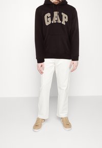 Felpa nera con cappuccio e logo "GAP" in beige, pantaloni grigi e scarpe color cammello. Tessuto morbido, tasca laterale sulla felpa, vestibilità casual.